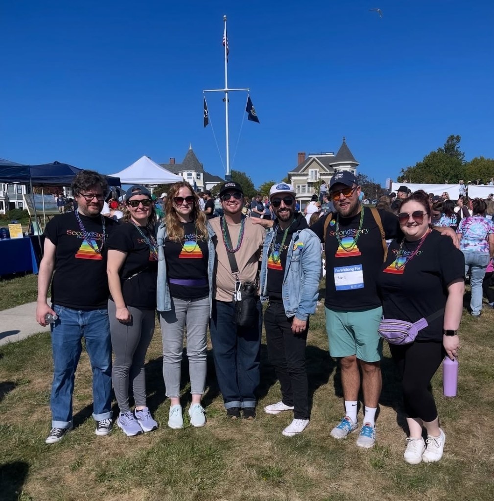 SecondStory company members stand at the eastern prom in Portland ME, in front of a flagpole. Behind them are several awnings and people. The SecondStory members are standing in a line with their arms around each other.