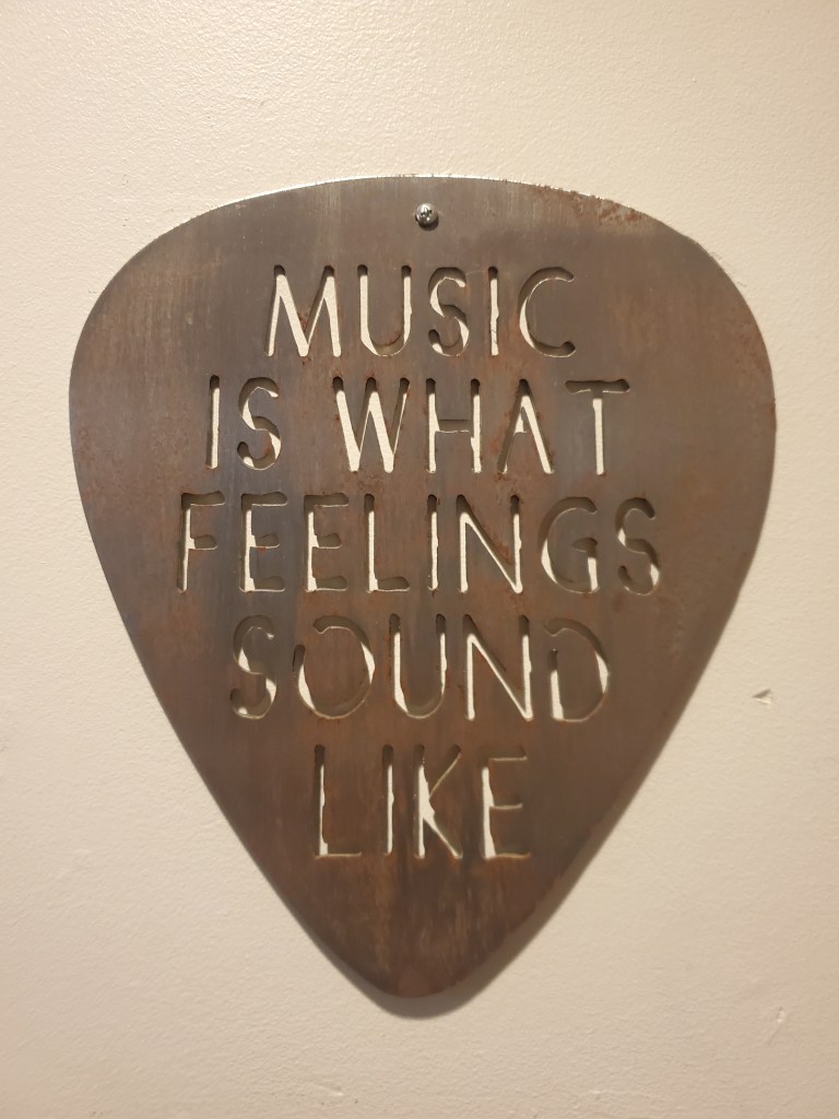 A large metal guitar pick hangs on the wall with the words "Music is what feelings sound like" cut out of it
