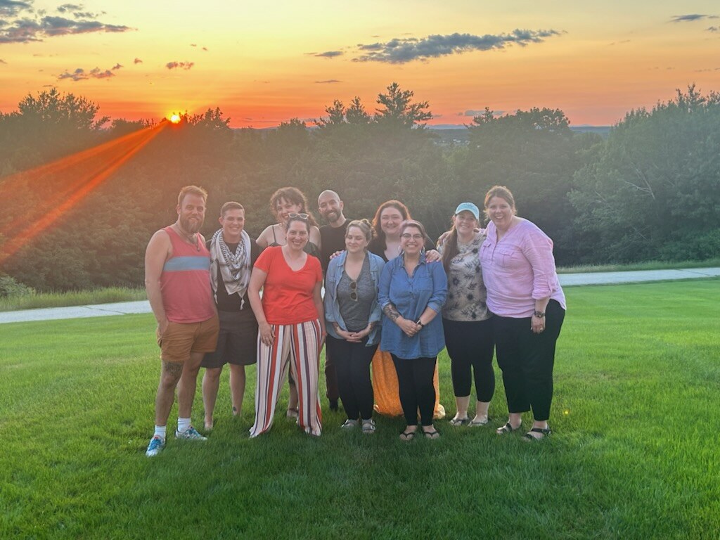 Company members stand in two lines with their arms around each other smiling. They are outside, standing on green grass. In the background are several leafy green trees and pine trees. The sun is setting behind them, the sky is orange, and there are a few clouds.