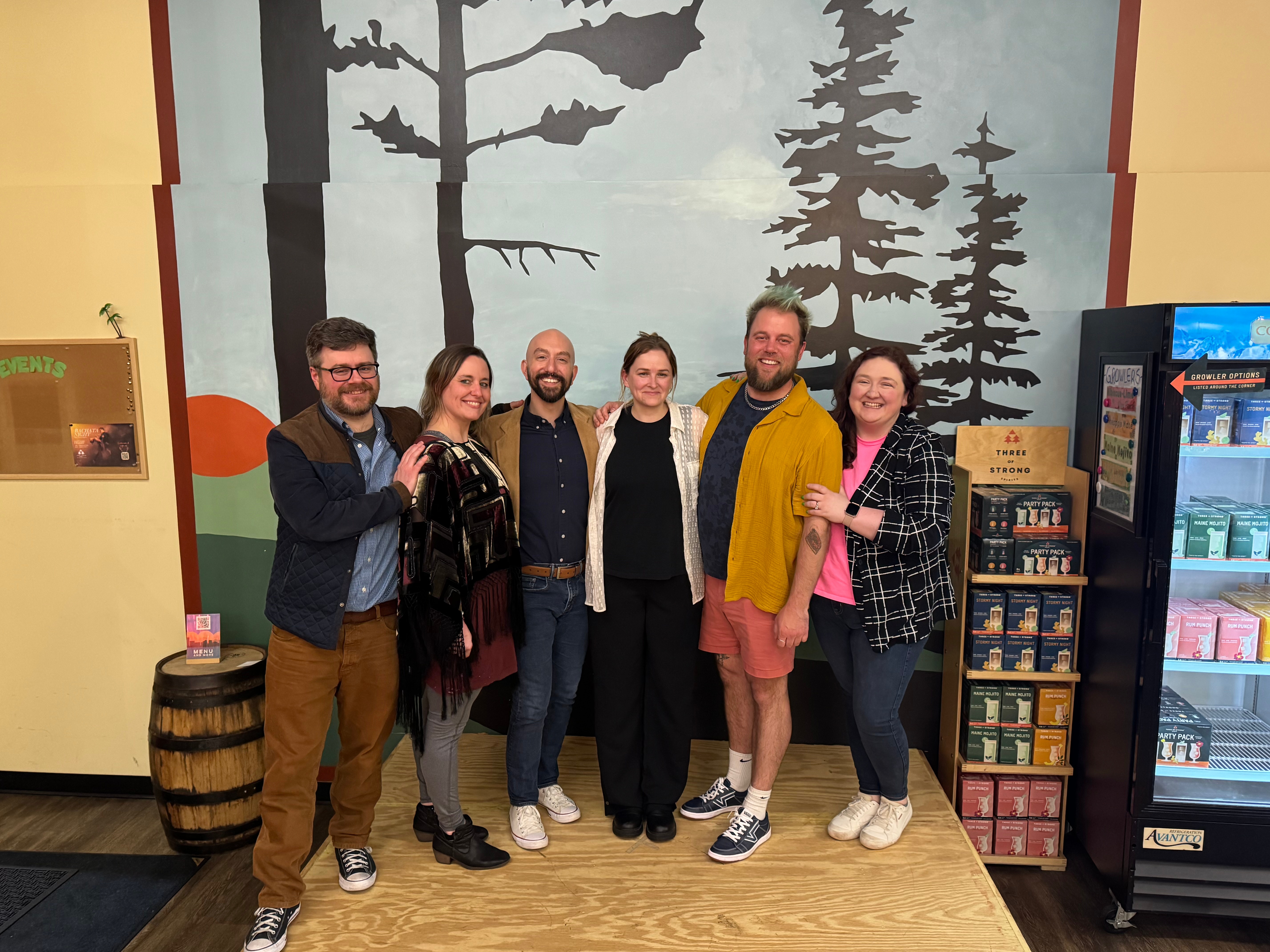6 of the 7 board members of SecondStory stand in a line on a stage with their arms around each other smiling. In the background is a mural with pine trees.