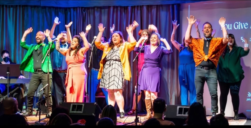 The company members are standing on stage during a group performance. They are in two lines and everyone is raising both hands in the air while singing. Everyone is wearing different, bright, primary colors.