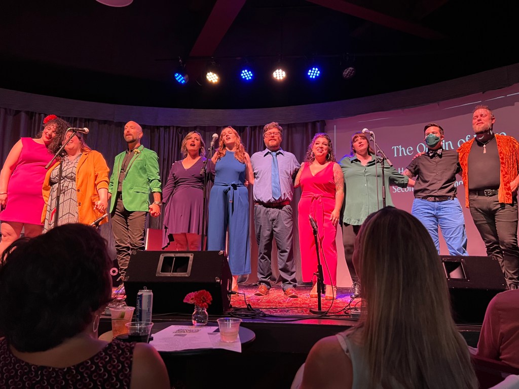 The cast of Love Is... stand on stage in a line with the arms around each other. They are all wearing bright solid colors.