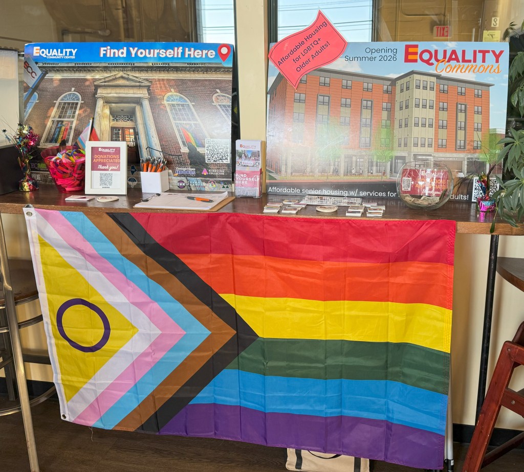 A table that has the intersex inclusive pride flag hanging in the front. On the table are two posters, one for the Equality Community Center and one for The Equality Commons. On the table are brochures, pins, stickers, and a sign up sheet for the ECC