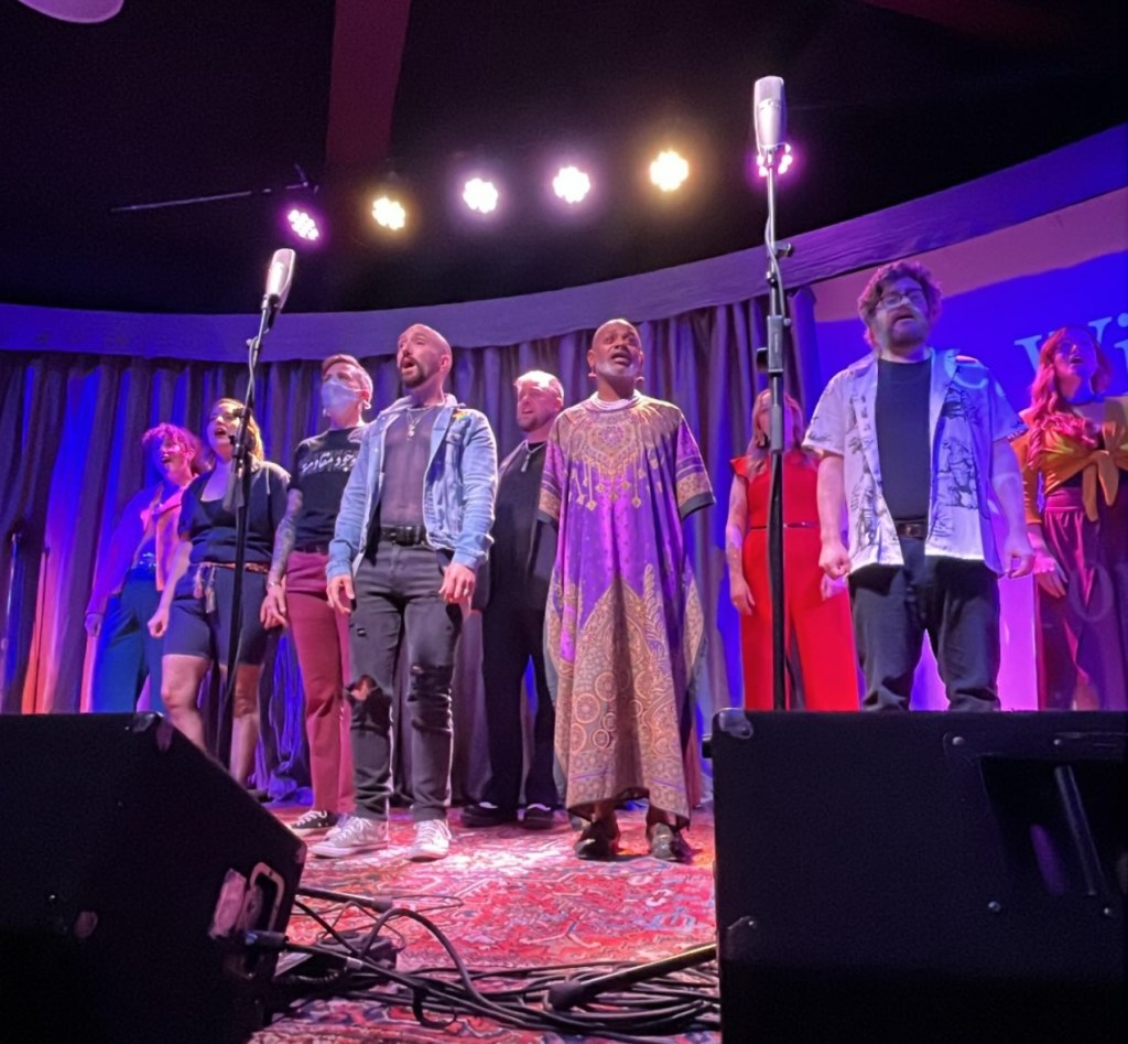 The SecondStory company is standing on stage, in a staggard line, wearing a variety of outfits. Everyone is singing.